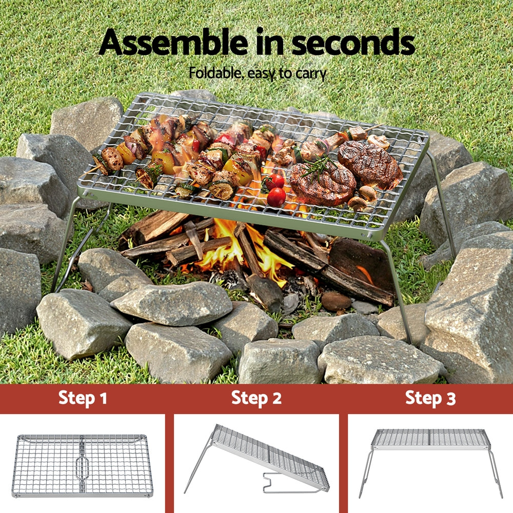 Portable grill with food over a campfire, surrounded by stones on grass